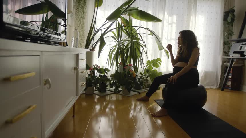 Pregnant woman with wavy hair black sports bra and leggings sits on black gym ball and eats fruit in the living room at home