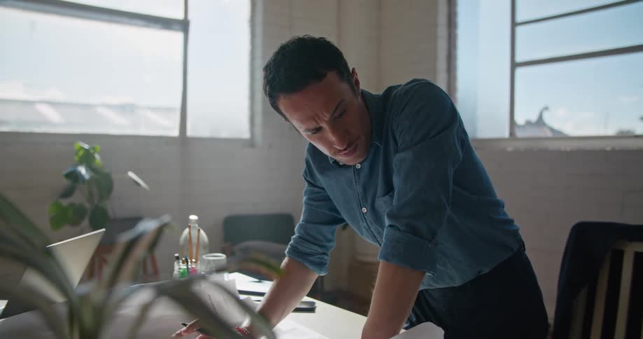A man is standing at his desk in an office while thinking and writing onto large blueprints during the day