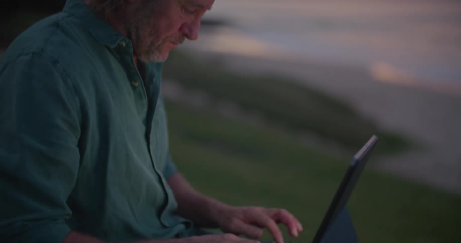 A man is typing on a laptop as he sits on a hill overlooking a beach