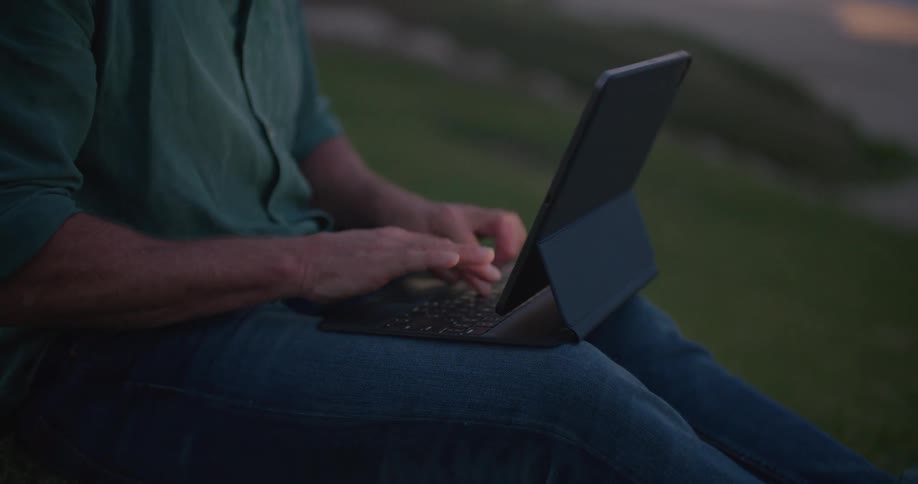 Man sits on grass near shore typing on computer with red tinted horizon and water in background