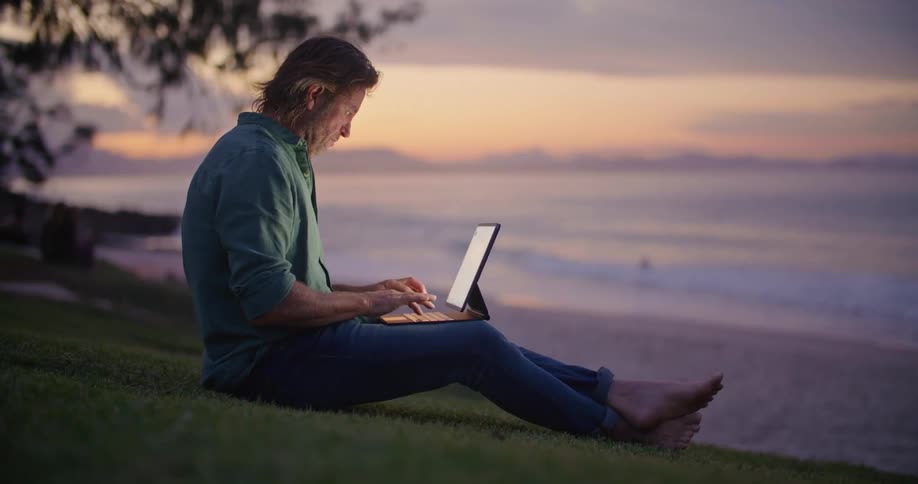 A man sits on the shore and types on a laptop