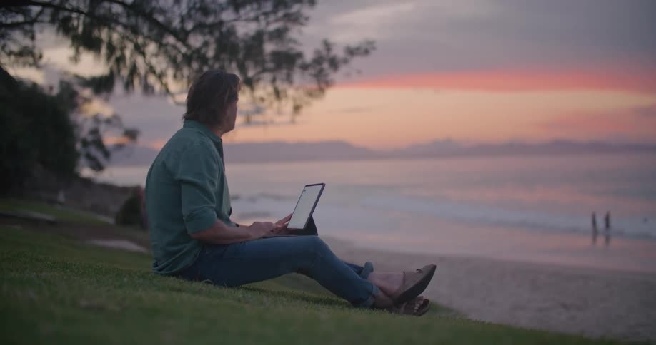 A man is sitting on a hill looking at a beach and typing on his laptop computer