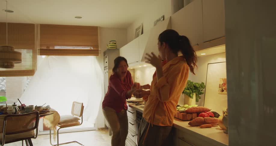 Friends stand in kitchen laughing as one throws and the other catches food