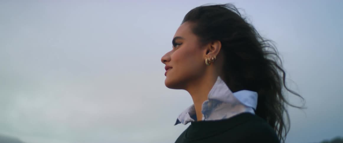 A woman walks along the shore and stretches her arms out wide and takes a deep breath as she looks towards the water