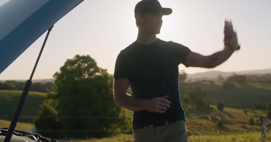 A man looks the trunk of a car and is looking at his phone as the sun shines brightly behind him