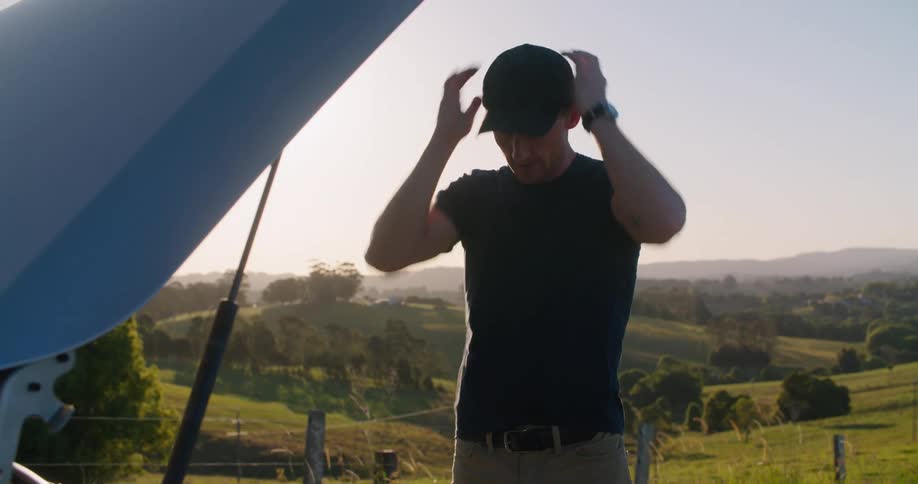 A man with a hat looking underneath the hood of the vehicle and a smart phone with the sky and clouds in the background