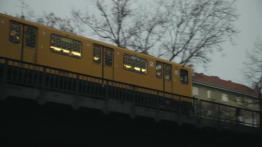Yellow train passes along bridge over street past apartment buildings and leafless trees under overcast sky
