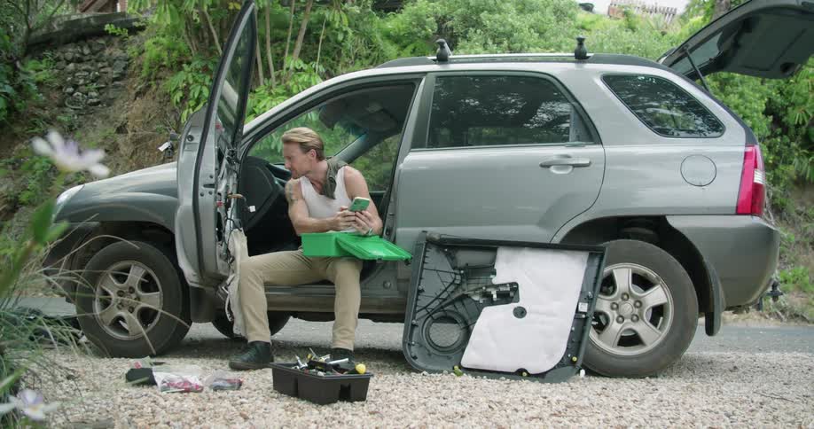 Young man sitting on the drivers seat of a car parked on a rural road uses his phone