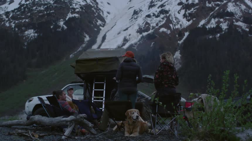 A group of friends sit around a grill below a mountain
