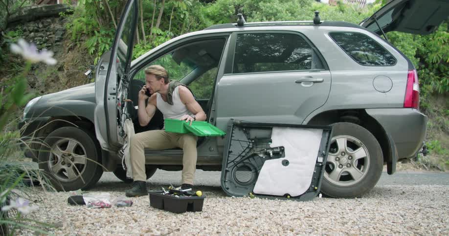 A man is sitting in his vehicle with the door open as he talks on the phone with the door panel removed and tools on the ground