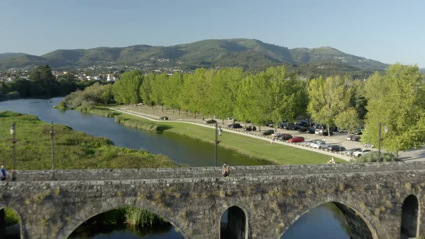 People walk along sidewalk paralleling river and passing by parking lot with town and mountain in distance