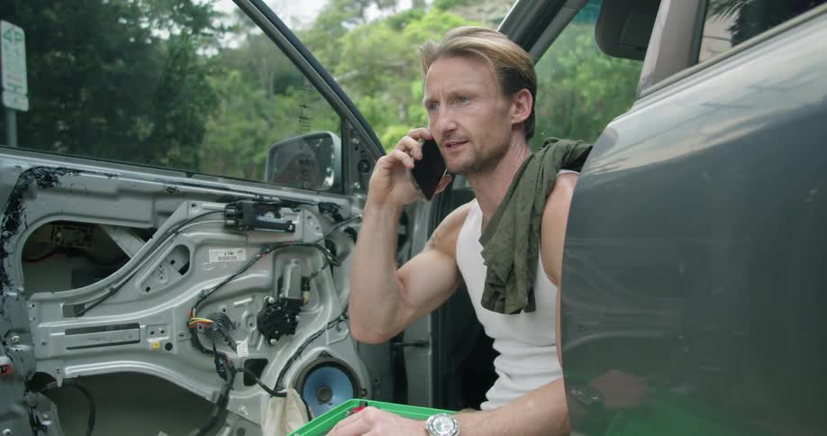 A man answers his phone while sitting in his car repairing a door panel