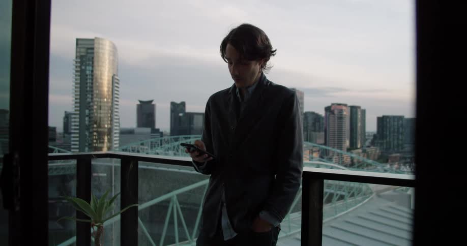 Man leans against balcony rail texting with city skyline in background