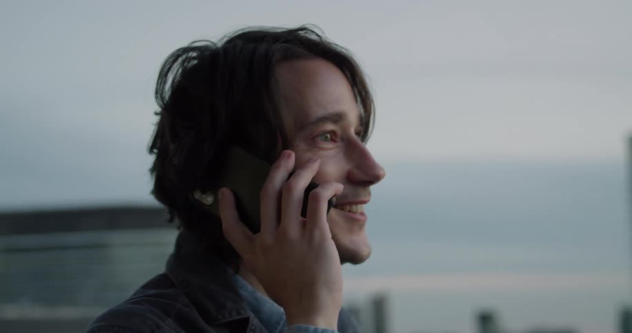 A man is outdoor talking and laughing on a cell phone in the city at dusk