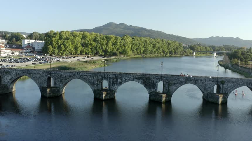 Boat floats near partly filled parking lot between arched stone bridge and another bridge on river leading toward mountains