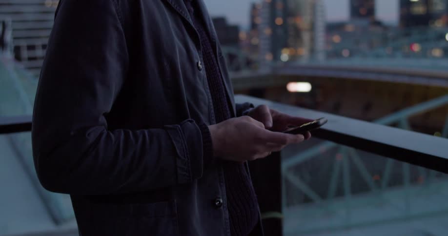 A man stands on a balcony and looks at the city skyline and then types on his cell phone