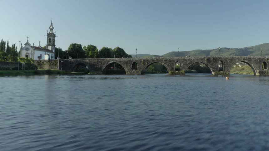Two people paddle boarding on a river with a bridge the sky and clouds in the background