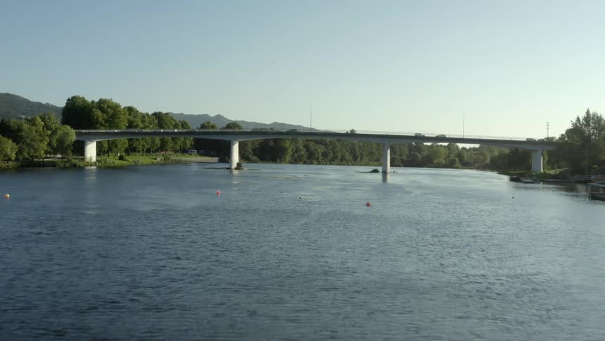 Traffic passesalong bridge over rippling river water near floating buoy with mountains and forest in background under a clear blue sky
