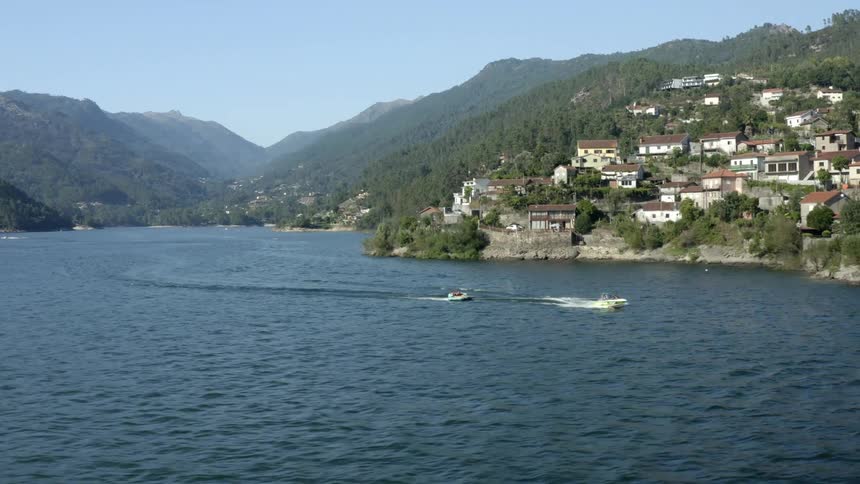 Aerial view of speedboat traveling sea passing by coastal town next to mountains with forest