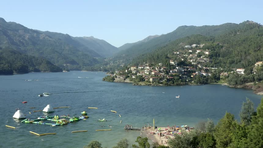 People are on a beach and boats are on a lake surrounded by mountains and houses along the side of the hill