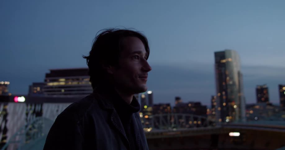 A man in a balcony using a smart phone with buildings the sky and clouds in the background