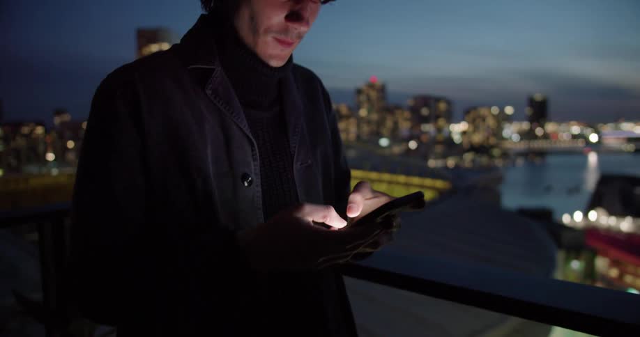 Man with black turtleneck sweater and coat using smartphone in dim lit office is silhouetted against the sky next to stadium at dusk