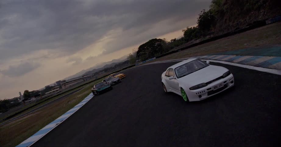 White car moves along race track with mountains and buildings in background under a cloudy overcast sky