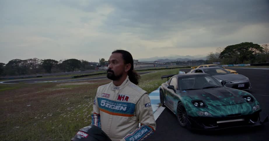 A race driver walks before cars parked on a race track while carrying his helmet on a cloudy evening