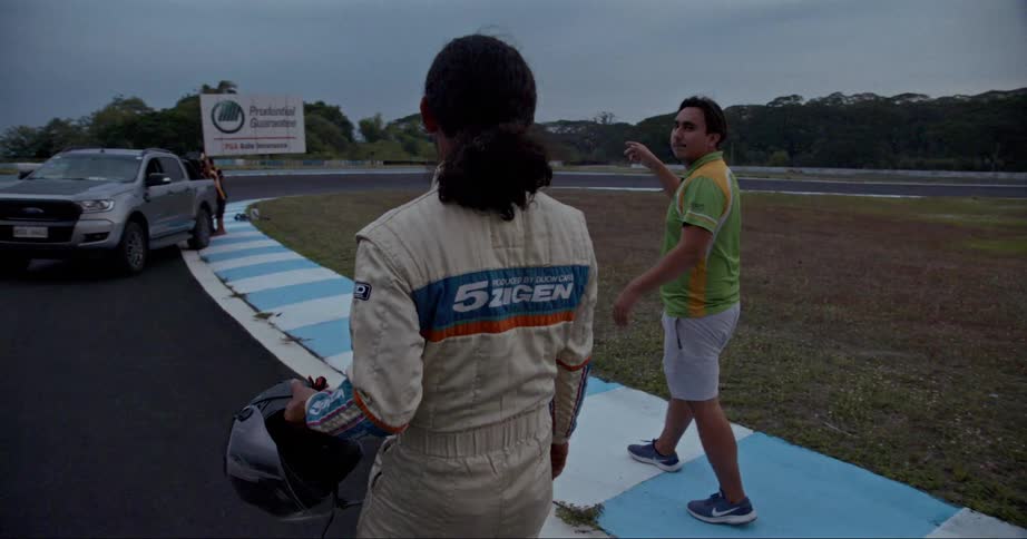 Two men walking on a racetrack with vehicles trees the sky and clouds in the background