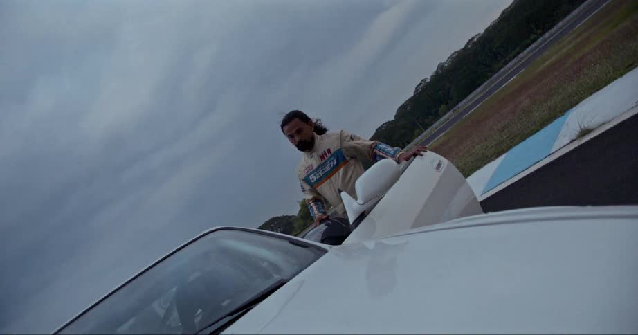 A race driver climbs into the drivers seat of a car on a race track on a cloudy evening