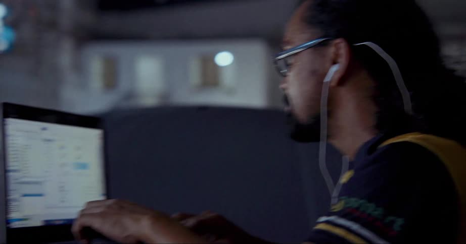 A man wearing earbuds types on a laptop resting atop the roof of a car outdoors at night
