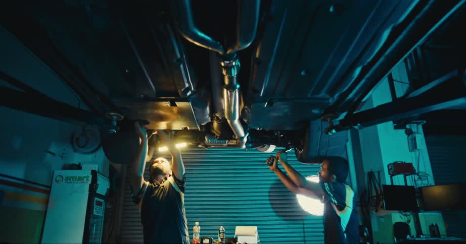 Two men are working on an elevated vehicle inside a large garage at night