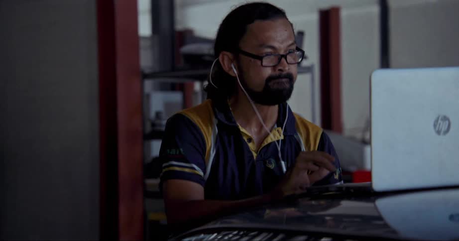 Man with long hair beard glasses and blue and yellow company shirt works on laptop when the coworkers are busy