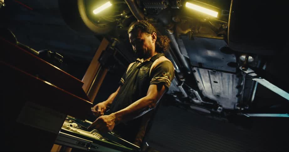 A mechanic standing underneath a hoisted up car picks up tools from a drawer