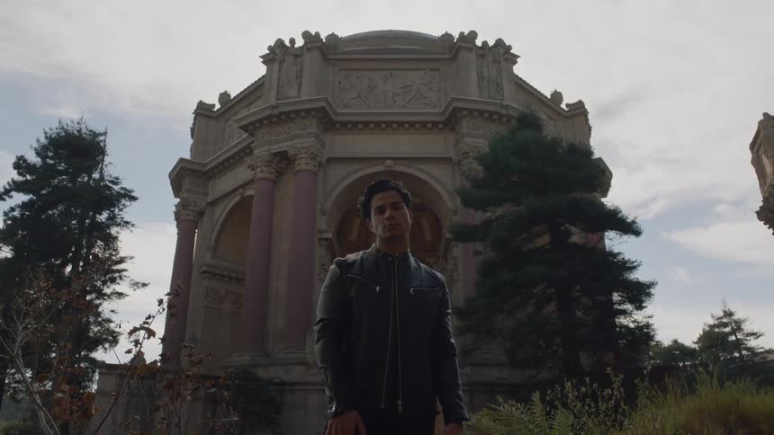 Man wearing leather jacket stands in garden near ornate detailed building under a cloudy sky