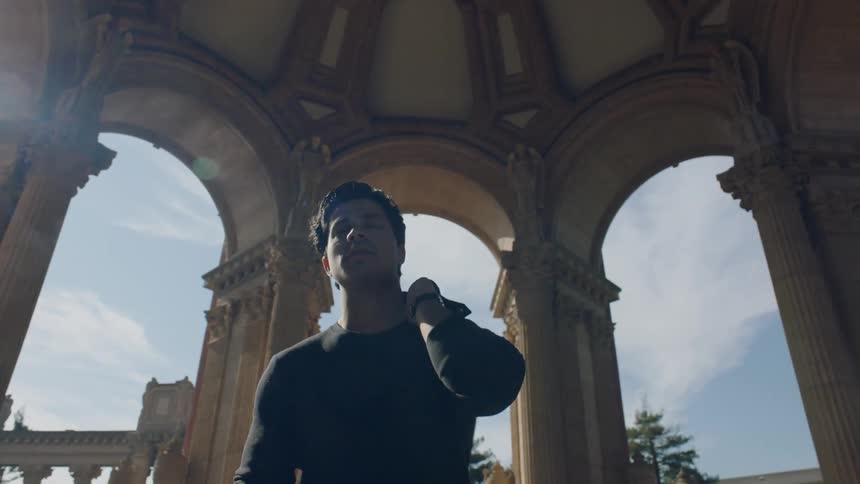 A man flips his jacket and holds it over his shoulder while standing under a columned dome on a sunny day