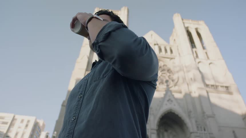 A man is standing in front of a cathedral while drinking a cup of coffee during the day
