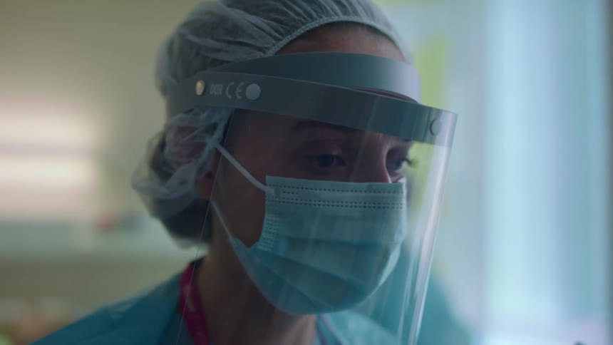 A nurse wearing a mask and face shield is waiting nervously near a patient in a hospital during the day