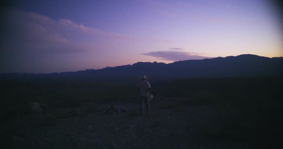 Man wearing a hat walking on the badlands near the mountains at evening