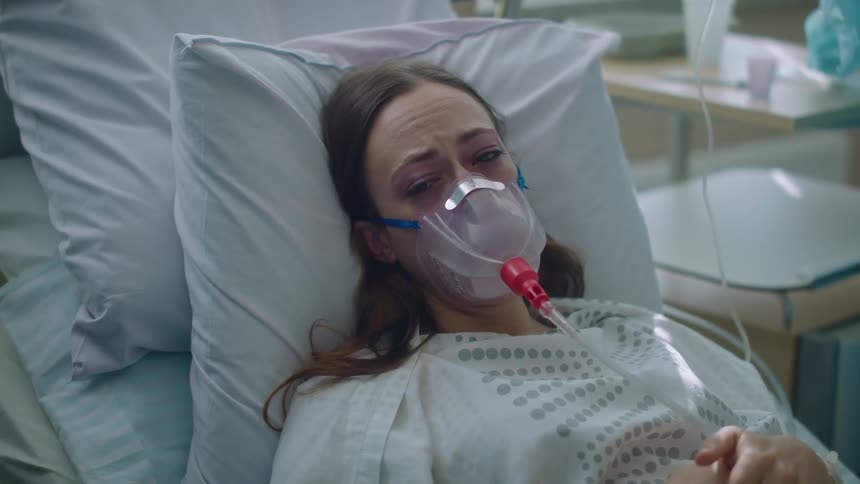 A woman wearing an oxygen mask is laying in a hospital bed with nurse working in the background
