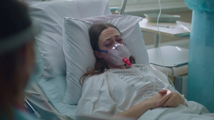 A woman in a hospital laying in a hospital bed with an oxygen mask with two doctors with scrubs
