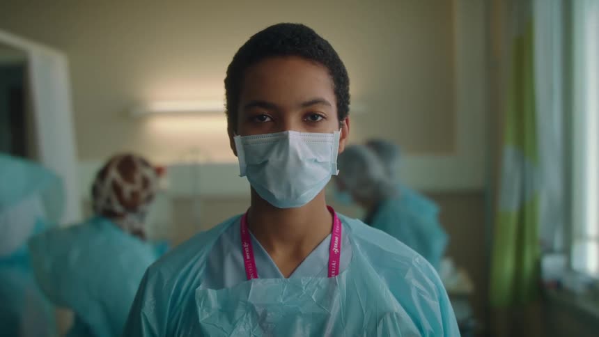 Female nurse with short hair mask and apron faces the camera in clinic