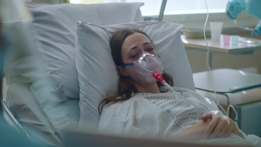 A woman in a hospital laying in a hospital bed with an oxygen mask with two doctors with scrubs