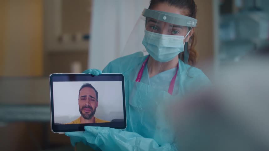 Female nurse with pony tail mask and scrub holds tablet with video call that man is crying