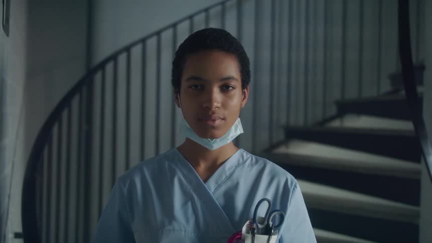 Nurse with short hair mask and scrubs faces the camera with eye contact in front of stairs