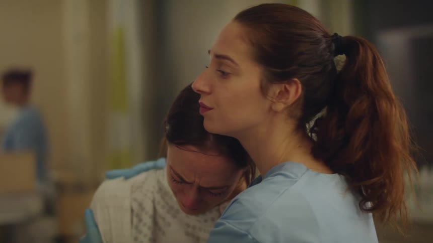 A woman wearing a hospital gown is crying and a nurse gives her a hug