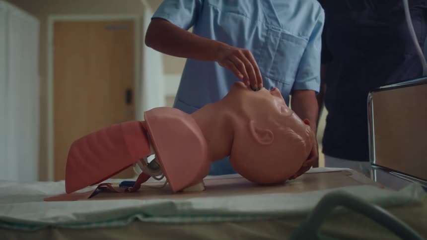 A nurse practicing on a dummy head in a hospital room with a wall and door in the background