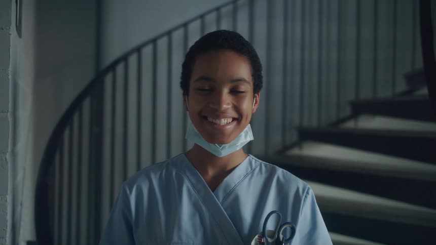 Female nurse with short hair mask and scrub faces the camera with eye contact smiling and standing in front of hospital stairs