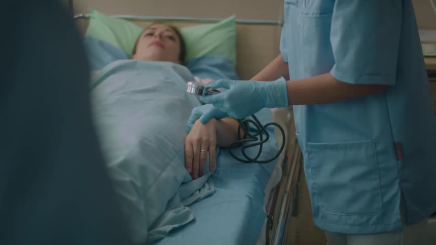 A woman in a hospital room laying in a hospital bed as a nurse with scrubs and gloves checks her pulse