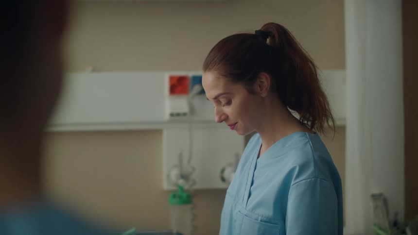 Young nurse wearing blue scrubs smiling inside a hospital room